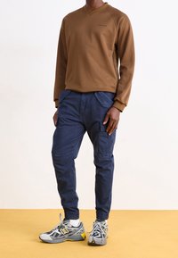 Brązowa bluza z długim rękawem i dekoltem w serek, w połączeniu z granatowymi spodniami typu cargo z bocznymi kieszeniami i elastycznymi mankietami, noszona z szarymi butami sportowymi.