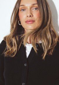 Melnā ribota kardigāna ar V-veida kaklu, kas piedāvā tumšus pogas. Fons ir vienkāršs balts, izceļot apģērba tekstūru un piegriezumu.