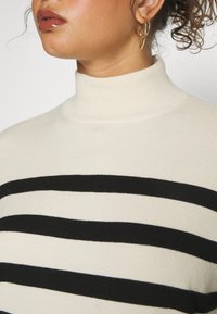 Beige poolokaulusneule, jossa paksut mustat raidat, pehmeä neulottu pinta, vartalonmyötäinen malli ja resorikaulus. Kultaiset renkaankorvakorut näkyvissä.