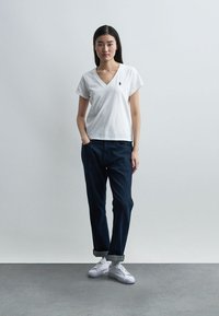 Γυναίκα που στέκεται μπροστά σε απλή λευκή τοιχοποιία, φορώντας λευκό T-shirt με V-λαιμόκοψη, σκούρο τζιν με μανσέτες και λευκά αθλητικά παπούτσια, με το ένα χέρι στην τσέπη.