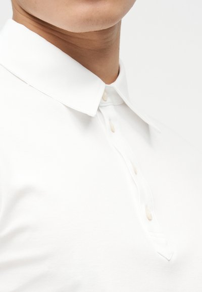 Κοντινό πλάνο ενός ατόμου που φοράει λευκό πουκάμισο με γιακά και μπεζ κουμπιά, δείχνοντας τον λαιμό και το πάνω μέρος του στήθους σε απλό φόντο.