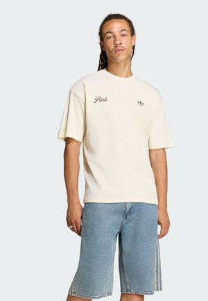 Giovane con capelli intrecciati che indossa una maglietta crema con il testo "Paris" e logo Adidas, abbinata a pantaloncini di denim azzurri.