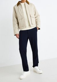 Giacca beige in cotone con colletto marrone, abbinata a pantaloni blu navy e sneakers bianche. La giacca presenta bottoni a pressione e due tasche frontali.