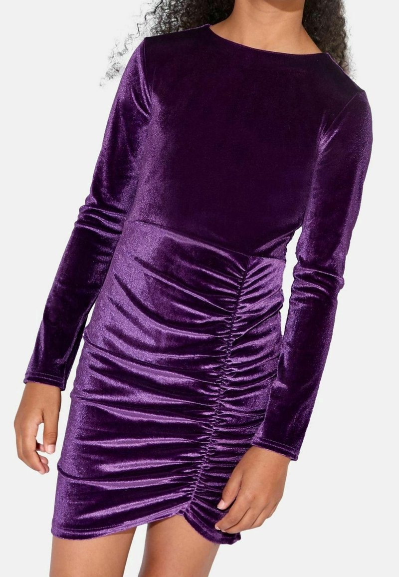 Fille portant une robe en velours violet à manches longues avec un détail froncé en diagonale sur le devant, sur un fond uni.