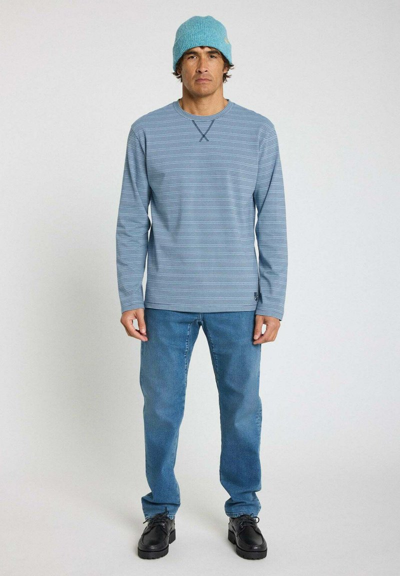 Chemise à rayures bleues à manches longues associée à un jean bleu clair et des chaussures noires ; un bonnet en tricot teal orne la tête, ajoutant texture et couleur.