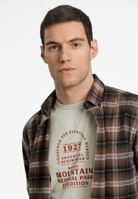 Braun kariertes Flanellhemd über einem hellgrünen T-Shirt mit rotem Text, das ein Grafikdesign zeigt, das mit Outdoor-Erkundung und Abenteuer zu tun hat.