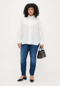 Hvit langermet skjorte med knapper, blå tettsittende jeans, svarte spisse flate sko, bærer en liten strukturert svart håndveske. Krøllete hår.