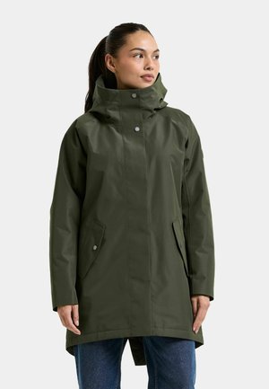 Femme portant un manteau imperméable à capuche vert foncé avec des fermetures à pression et des poches avant, associé à un jean bleu.