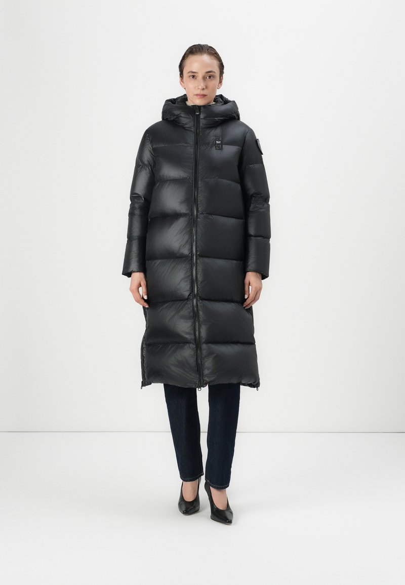 Lang zwarte pufferjas met een capuchon, met quilted delen, een rits aan de voorkant en zijzakken. Gedragen over donkere jeans en zwarte schoenen.