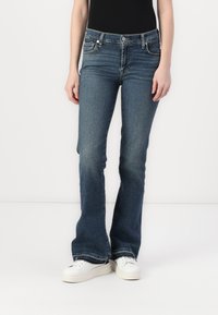 Jeans evazați albaștri din denim elastic, cu talie medie, cinci buzunare și o ușoară decolorare. Purtați cu adidași albi.