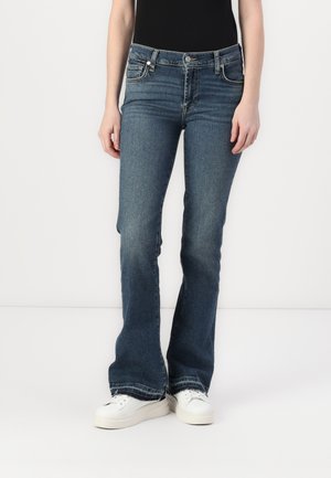 Blue flared jeans in stretchy denim, featuring a mid-rise waist, five pockets, and subtle whiskering. Worn with white sneakers.