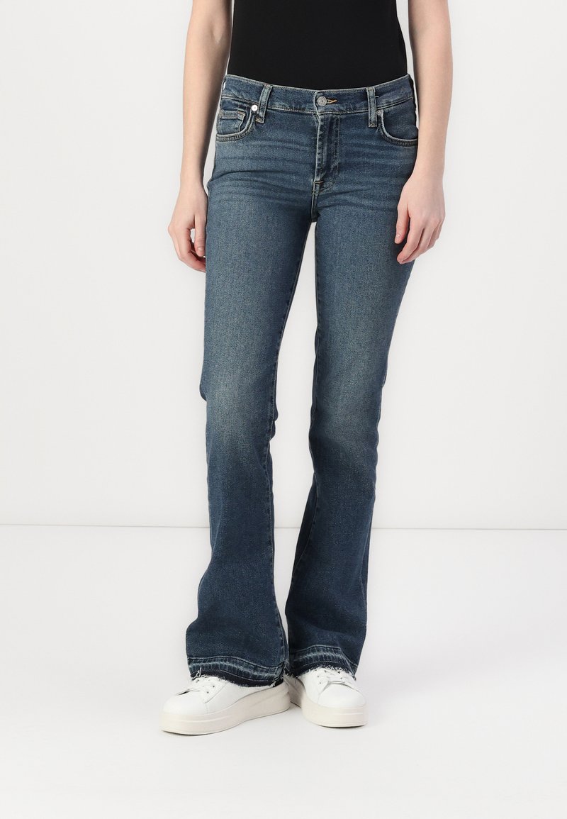 Jeans evazați albaștri din denim elastic, cu talie medie, cinci buzunare și o ușoară decolorare. Purtați cu adidași albi.