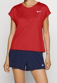 Punainen urheilupaita lyhyillä hihoilla, teksturoitua kangasta ja valkoinen Nike-logo; yhdistetty tumma sininen shortsien kanssa.