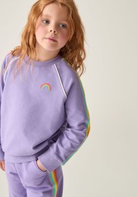 Lila kapucnis pulóver szivárványcsíkos díszítéssel, amelyen egy kis hímzett szivárvány található a mellkasi részen. Puha anyag, laza szabással.