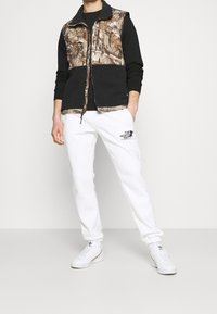 Bezrękawnik w wzór moro z czarnym polarowym dołem, w połączeniu z białymi dresami z czarnym logiem. Całość dopełniają białe sneakersy.