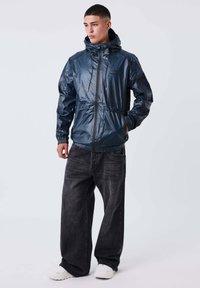 Giovane uomo che indossa una giacca a vento lucida di colore blu navy con cappuccio, jeans neri larghi e sneakers bianche, in piedi con una mano in tasca contro uno sfondo semplice.