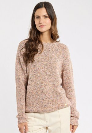 Woman with long wavy hair wearing a multicolored knit sweater and cream pants, standing against a plain white background.