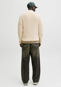 Pull en maille crème avec texture côtelée, coupe oversize. Porté avec un jean large à dégradé sombre. Casquette de couleur vert foncé avec une visière plate.