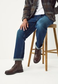 Braune Leder-Schnürstiefel mit glatter Oberfläche, die auf einem Holzstuhl stehen. Kombiniert mit blauen Jeans und einem karierten Flanellhemd.