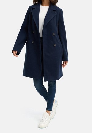 Femme portant un manteau bleu foncé à double boutonnage, une chemise blanche, un jean skinny foncé et des baskets blanches, debout avec une jambe croisée.
