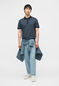 Πόλο πουκάμισο με λωρίδες σε ναυτικό μπλε και γκρι, σε συνδυασμό με ανοιχτό μπλε παντελόνι με λεπτές ρίγες και τζιν μπουφάν δεμένο γύρω από τη μέση. Λευκά αθλητικά παπούτσια.