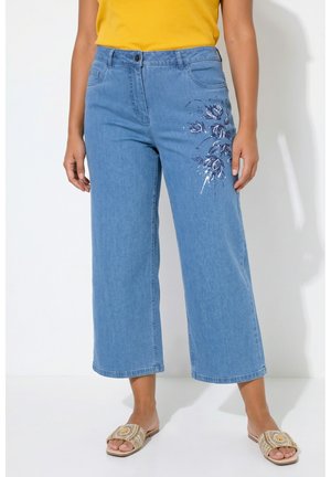 Kvinde iført lyseblå vidde kortere jeans med mørkt blomsterbroderi på låret og beige åbne sandaler.