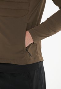 Brown long-sleeve top with a smooth texture, featuring a zippered pocket on the side and a slightly elastic bottom hem.