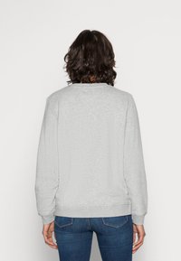 Felpa grigia con collo rotondo, polsini e orlo a costine, realizzata in un tessuto testurizzato. Indossata con jeans blu. Vista da dietro.