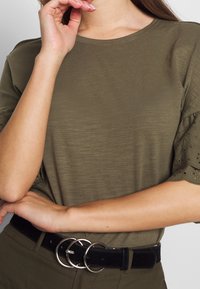 Femme portant un haut olive avec des manches en dentelle à l'œillet et une ceinture noire avec une boucle double en argent, les bras croisés à la taille.