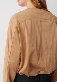 Top in suede color nocciola con una vestibilità ampia, scollatura rotonda e dettagli di cucitura in evidenza. Il tessuto ha una consistenza morbida e opaca.