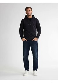 Sudadera negra con capucha y cordones, bolsillo frontal y logo en el pecho, combinada con vaqueros azul oscuro y zapatillas blancas sobre un fondo neutro.