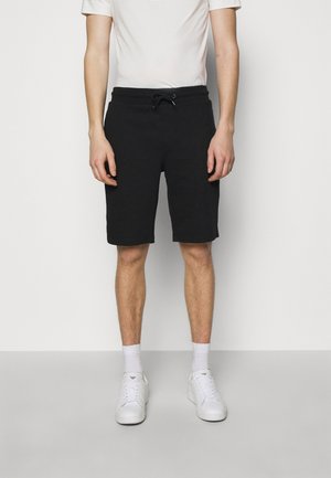 Man draagt zwarte shorts met aantrekkoord, wit T-shirt met korte mouwen, witte sokken en witte sneakers, staand tegen een effen achtergrond.
