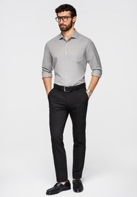 Homme avec barbe et lunettes portant une chemise gris clair aux manches retroussées, un pantalon noir, une ceinture noire, une montre au poignet et des chaussures noires, debout les mains dans les poches.