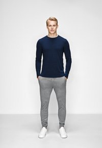 Mørkeblå langærmet skjorte med tekstureret overflade, parret med lysegrå sweatpants og hvide sneakers, stående foran en ensfarvet baggrund.