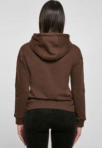 Sudadera marrón con una gran capucha, hombros caídos y puños acanalados. Llevada sobre pantalones negros texturizados, vista desde la parte posterior.