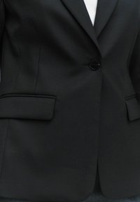 Svart skräddarsydd blazer med en enda svart knapp, smala slag och två framfickor. Slät, strukturerad tyg med en matt yta.