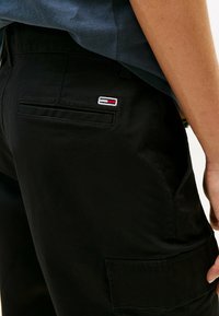 Pantalones cargo negros con una textura suave, que cuentan con un bolsillo trasero y un pequeño logo bordado en rojo, blanco y azul.