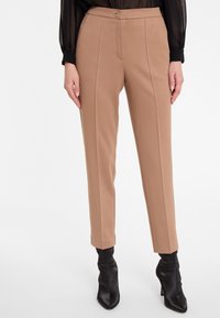 Pantalons beiges ajustés avec une jambe droite, dotés d'une fermeture à bouton sur le devant et de détails de couture verticaux. Portés avec des bottes noires à talons.