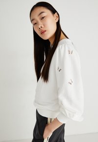 Sudadera blanca con mangas abullonadas, que presenta un detalle de mariposa recortado. Tejido suave y puños de canalé, combinada con pantalones negros.