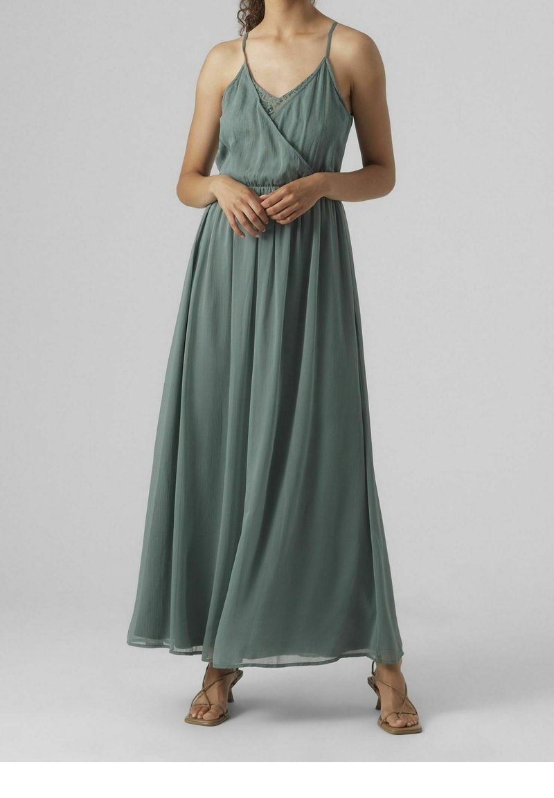 Femme portant une robe verte sans manches, longue jusqu'au sol, avec un galon de dentelle et de fines bretelles, associée à des talons beiges à brides.