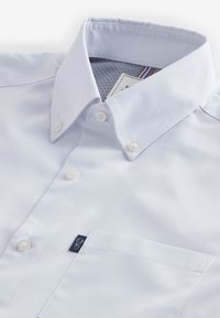 Chemise blanche à boutons avec col pointu, poche poitrine et petite étiquette sombre sur le bord de la poche.