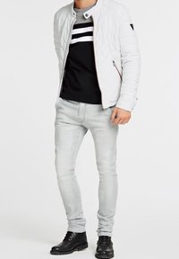 Veste matelassée blanche à col haut, portée par-dessus un haut noir avec des rayures blanches. Jeans slim gris clair et bottes noires complètent la tenue.