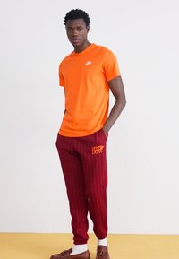 Orange Nike t-shirt with a crew neckline, paired with burgundy pinstriped pants featuring "Just Do It" in orange. Model stands against a plain background.