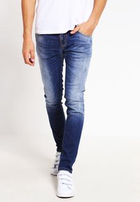 Smala blå jeans, urtvättade vid låren; gjorda av denim med en slät textur, kombinerat med vita sneakers, visade i en gående pose.