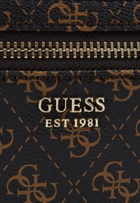 Matériau synthétique noir avec un motif de logo marron. Quincaillerie dorée affichant "GUESS" et "EST 1981". Fermeture zippée.