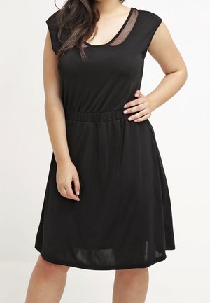 Nainen, joka pukeutunut hihattomaan mustaan mekkoon, jossa on verkkokuvioinen pääntie, joustava vyötärö ja polvenpituinen helma.