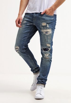 Man wearing blue distressed jeans with multiple rips and white sneakers, standing with one hand in pocket against plain background.