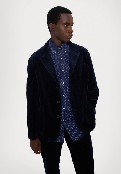 Marineblå kordfløyelsblazer med brune knapper, parret med en blå prikkete skjorte. Glatt tekstur og skreddersydd passform. To sidelommer.