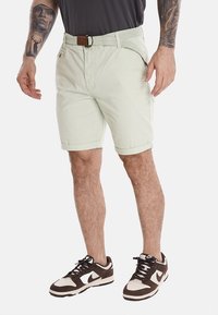 Pantaloni shorts verde chiaro in cotone, con una cintura intrecciata, tasche frontali e risvolti. Abbinati a sneakers marroni e bianche.