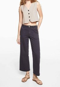 Béžová zkrácená vesta s vertikálními pruhy a černými knoflíky, ve spojení s tmavě šedými širokými kalhotami a sandály na ploché podrážce. Hladká textura látky.